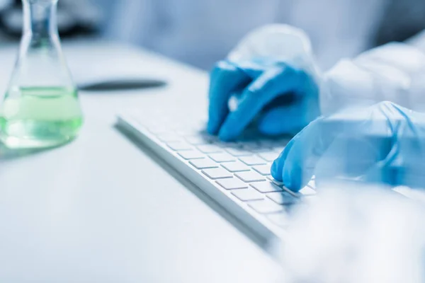 Partial view of scientist typing on computer keyboard near blurred flask — Stock Photo