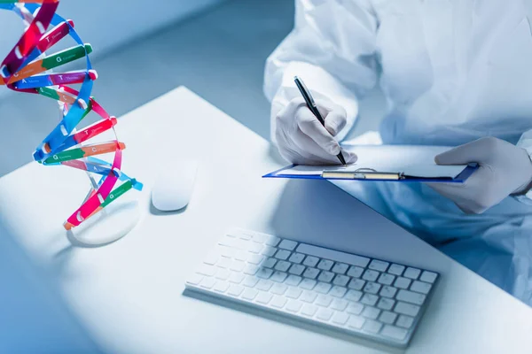 Abgeschnittene Ansicht des Wissenschaftlers, der auf Klemmbrett neben dna-Modell und Computertastatur schreibt — Stockfoto