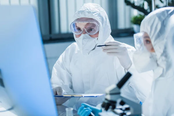 Virologe im Warnanzug hält Klemmbrett in der Hand und zeigt auf verschwommenen Monitor in der Nähe des Kollegen — Stockfoto