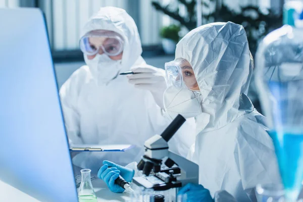 Mujer en traje de materiales peligrosos que trabaja con microscopio cerca del monitor de la computadora y colega borrosa - foto de stock