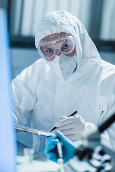 Virólogo en traje de materiales peligrosos y gafas mirando a la cámara en primer plano borroso - foto de stock