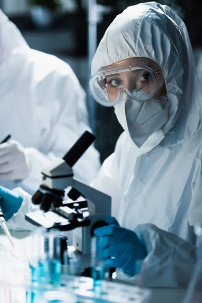 Femme en combinaison Hazmat, lunettes et masque médical regardant la caméra près du microscope — Photo de stock