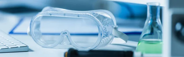 Transparent goggles near blurred flask in laboratory, banner — Stock Photo