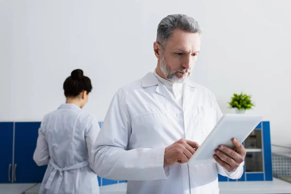 Científico barbudo de capa blanca usando tableta digital cerca de colega en laboratorio - foto de stock