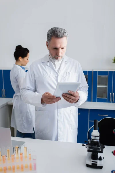 Wissenschaftler mittleren Alters betrachten digitales Tablet, während sie in der Nähe von Kollegen im Labor arbeiten — Stockfoto