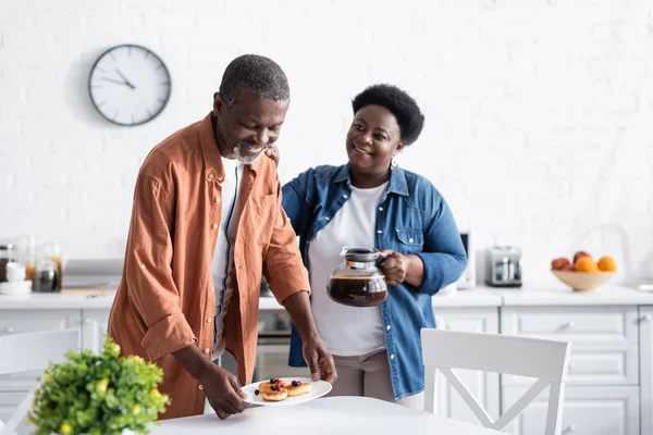 Feliz homem americano africano segurando panquecas na placa perto da esposa com panela de café — Fotografia de Stock