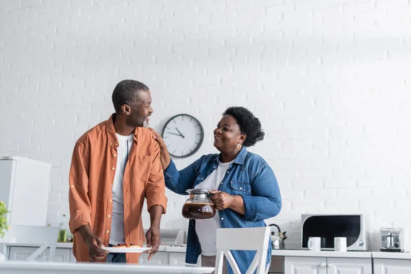 Felice uomo africano americano che tiene frittelle sul piatto e guardando moglie con caffettiera — Foto stock