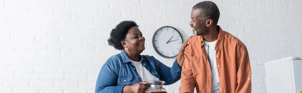 Hombre afroamericano feliz mirando sonriente esposa con cafetera, bandera - foto de stock