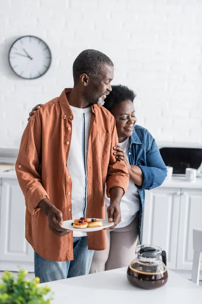 Heureuse femme afro-américaine étreignant mari tenant des crêpes dans la cuisine — Photo de stock