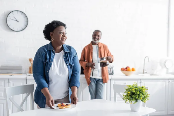 Feliz mulher americana africana sênior segurando placa com panquecas perto marido borrado na cozinha — Fotografia de Stock