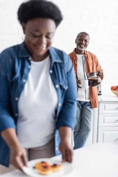 Glücklich Senior afrikanisch-amerikanischer Mann mit Kaffeekanne und Blick auf verschwommene Frau — Stockfoto