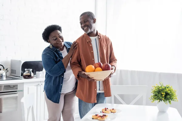 Anziano uomo africano americano che tiene ciotola con frutta fresca vicino sorridente moglie in cucina — Foto stock