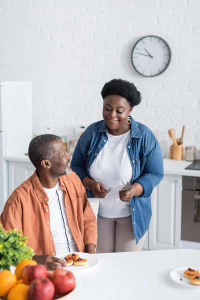 Mujer afroamericana feliz y mayor sosteniendo cubiertos cerca del marido durante el desayuno - foto de stock