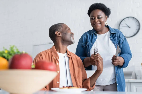 Feliz y mayor afroamericana mujer sosteniendo cubiertos cerca sonriente marido durante el desayuno - foto de stock