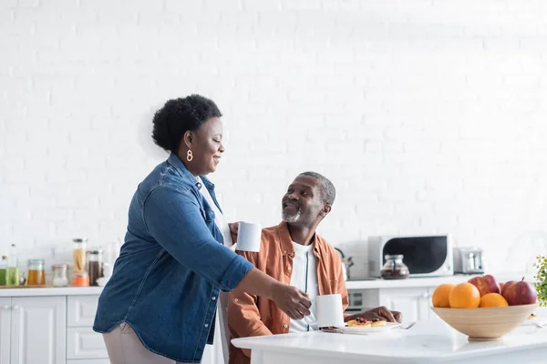 Счастливая африканская американка держит чашки кофе возле улыбающегося мужа во время завтрака — стоковое фото