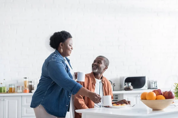 Mujer afroamericana feliz sosteniendo tazas de café cerca de marido sonriente durante el desayuno - foto de stock