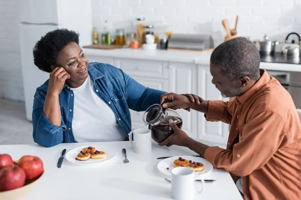 Homem americano africano sênior derramando café para esposa alegre durante o café da manhã — Fotografia de Stock