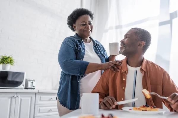 Sénior afroamericano hombre desayunando y mirando feliz esposa - foto de stock
