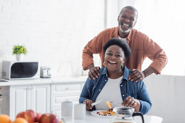 Feliz e sênior africano americano homem de pé atrás da esposa durante o café da manhã — Fotografia de Stock