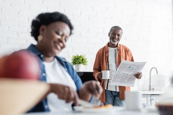 Glücklicher und älterer afrikanisch-amerikanischer Mann hält Tasse und Zeitung in der Hand, während er die verschwommene Frau im Vordergrund betrachtet — Stockfoto