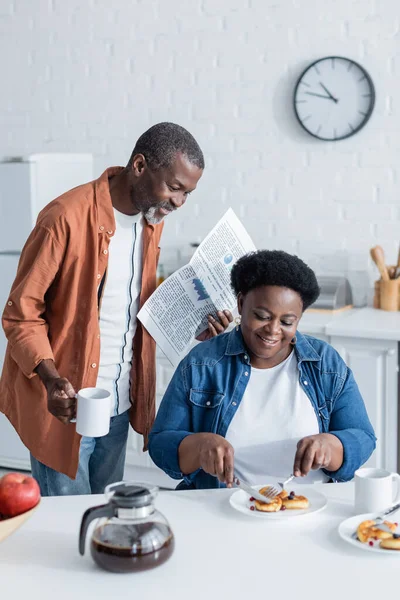 Glücklicher und älterer afrikanisch-amerikanischer Mann hält Tasse und Zeitung in der Hand, während er seiner Frau beim Frühstück zusieht — Stockfoto