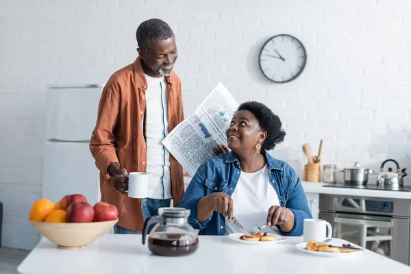 Homem americano africano alegre e sênior que prende o copo e o jornal ao olhar na esposa que toma o café da manhã — Fotografia de Stock