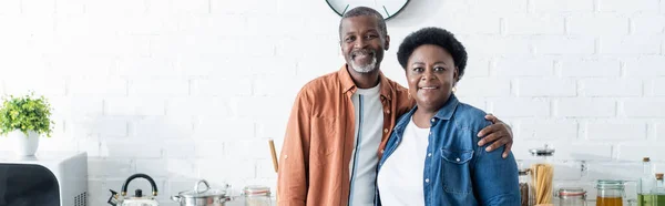 Happy senior african american couple looking at camera in kitchen, banner — Stock Photo