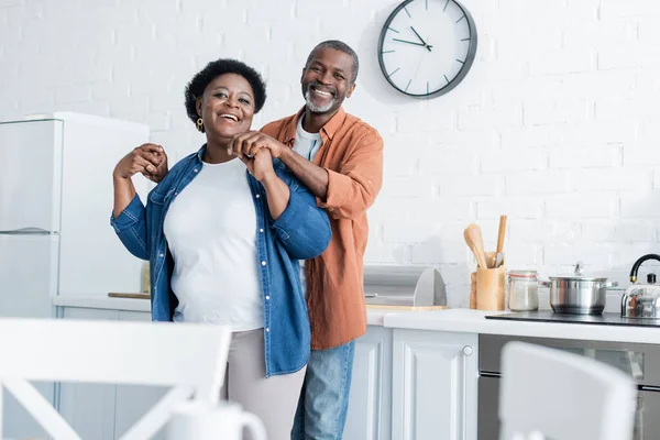 Happy senior african american man holding hands of smiling wife in kitchen — Stock Photo