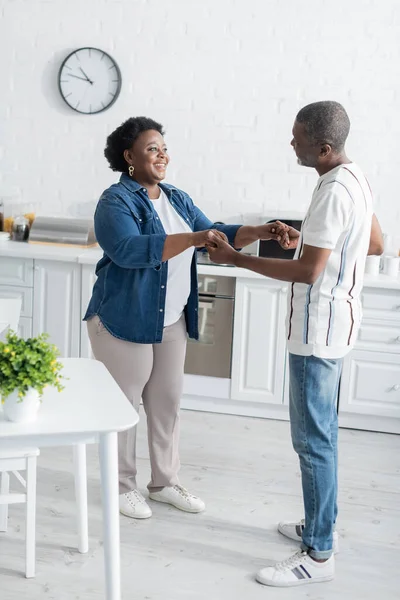 Full length of happy senior african american man holding hands with smiling wife while dancing in kitchen — Stock Photo