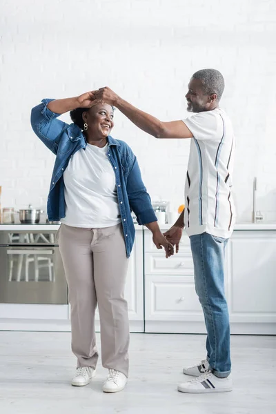 Volle Länge von fröhlichen Senior afrikanisch-amerikanischen Mann Händchen haltend mit lächelnder Frau beim Tanzen in der Küche — Stockfoto