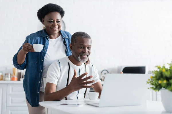 Sénior afroamericano hombre gesto cerca de la esposa mientras mira el ordenador portátil durante el chat de vídeo - foto de stock