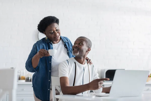 Barbudo afroamericano hombre apuntando a la computadora portátil y mirando a la esposa - foto de stock