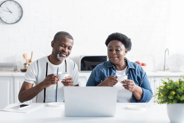 Sênior afro-americano marido e mulher segurando copos e assistindo filme no laptop — Fotografia de Stock