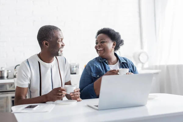 Retired african american husband and wife holding cups and looking at each other near laptop — Stock Photo