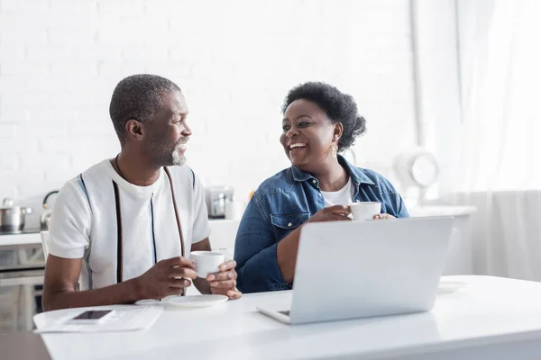 Retirado afroamericano marido y esposa sosteniendo tazas y mirando unos a otros cerca de la computadora portátil - foto de stock