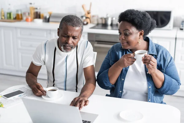 Retirado afroamericano marido y esposa sosteniendo tazas y viendo la película en el ordenador portátil - foto de stock