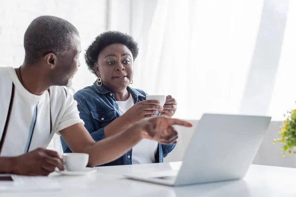 Afrikanischer Rentner zeigt mit Tasse auf Laptop neben Ehefrau — Stockfoto