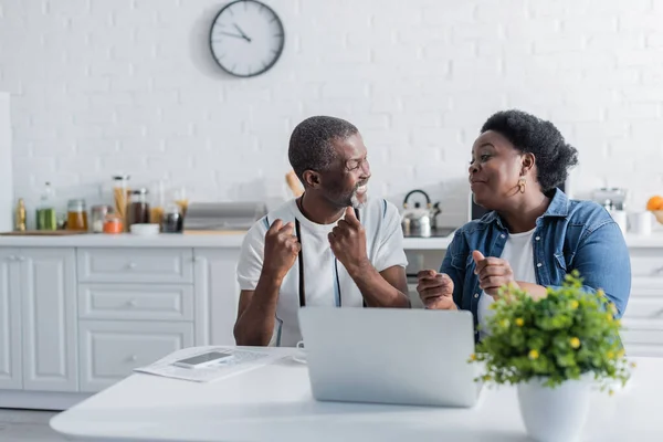 Marido y esposa afroamericanos mayores regocijándose y mirándose cerca de la computadora portátil - foto de stock