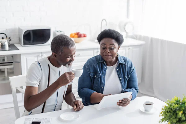 Mulher americana africana sênior mostrando tablet digital para marido com xícara de café — Fotografia de Stock