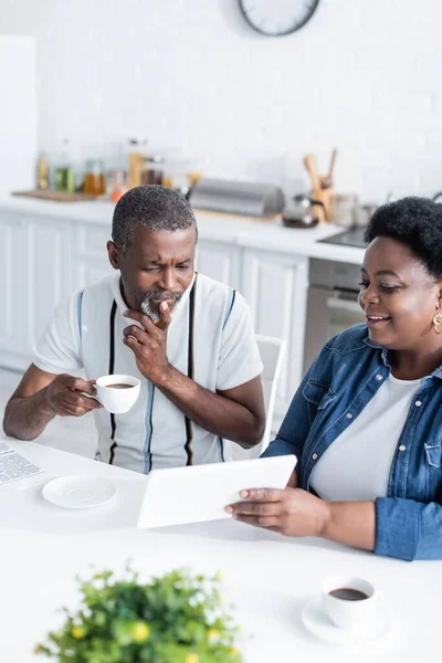 Alegre mujer afroamericana mostrando tableta digital a marido mayor con taza de café - foto de stock
