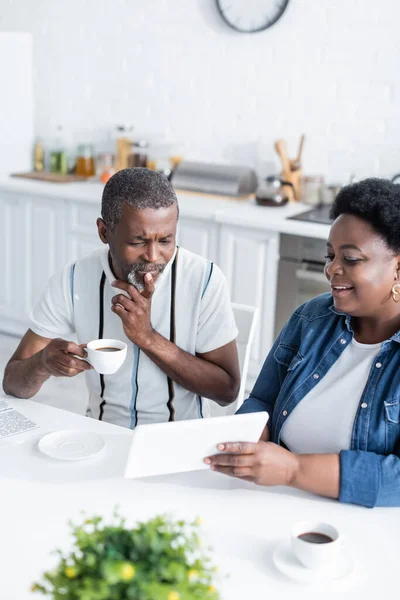 Mulher americana africana alegre mostrando tablet digital para marido sênior com xícara de café — Fotografia de Stock