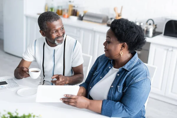 Cheerful african american woman showing digital tablet to smiling husband with cup of coffee — Stock Photo