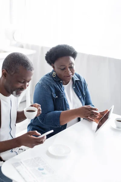 Donna afroamericana in possesso di tablet digitale vicino marito allegro con tazza di caffè utilizzando smartphone — Foto stock