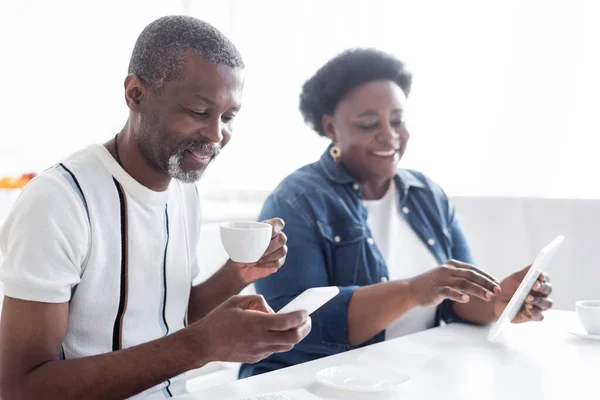 Homem americano africano sênior com xícara de café usando smartphone perto da esposa borrada com tablet digital — Fotografia de Stock