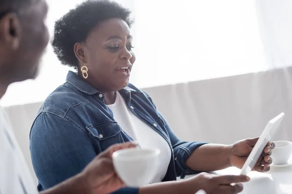 Senior afro-américaine femme tenant tablette numérique près mari flou avec tasse de café — Photo de stock