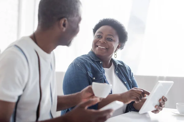 Sonriente mujer afroamericana apuntando a la tableta digital y mirando al marido mayor con taza de café - foto de stock