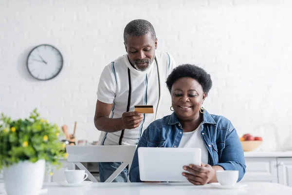 Senior africano americano hombre celebración de tarjeta de crédito mientras que las compras en línea con feliz esposa - foto de stock