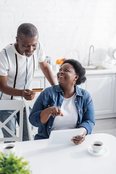 Senior africano americano hombre celebración de tarjeta de crédito mientras que las compras en línea cerca feliz esposa apuntando a la tableta digital - foto de stock