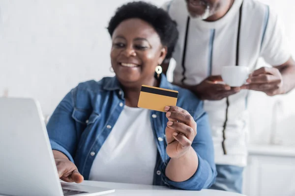 Mujer afroamericana mayor sosteniendo tarjeta de crédito cerca de la computadora portátil y esposo borroso con taza - foto de stock