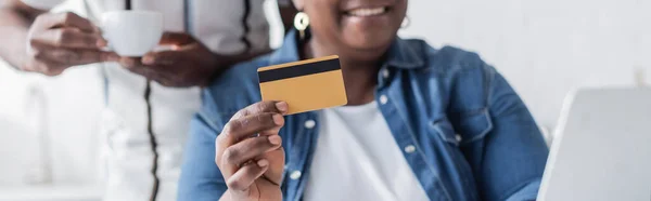 Vista cortada da mulher americana africana sênior segurando cartão de crédito perto de laptop e marido borrado com copo, banner — Fotografia de Stock